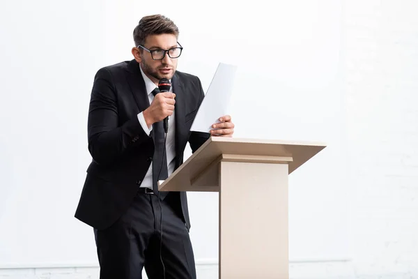 Испуганный бизнесмен в костюме, стоящий на трибуне и выступающий во время конференции изолированный по белому — стоковое фото