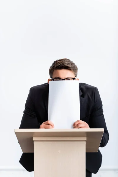 Scared businessman in suit standing at podium tribune and obscuring face with paper during conference isolated on white — Stock Photo