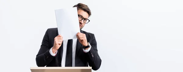 Panoramaaufnahme eines verängstigten Geschäftsmannes im Anzug, der auf der Tribüne steht und sein Gesicht während der Konferenz mit Papier verdeckt, isoliert auf Weiß — Stockfoto