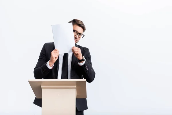 Scared businessman in suit standing at podium tribune and obscuring face with paper during conference isolated on white — Stock Photo