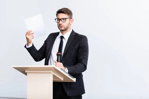 Hombre de negocios asustado en traje de pie en tribuna podio y la celebración de micrófono durante la conferencia aislado en blanco - foto de stock