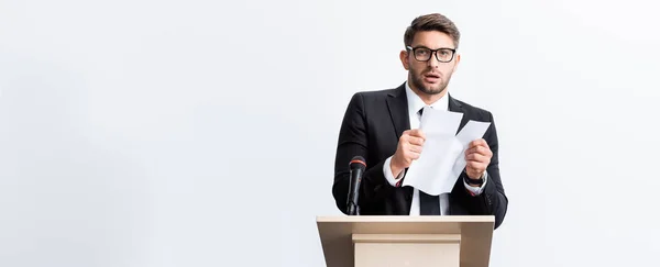 Panoramic shot of scared businessman in suit standing at podium tribune and rearing paper during conference isolated on white — Stock Photo