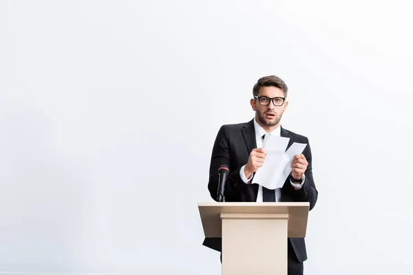 Asustado hombre de negocios en traje de pie en tribuna podio y crianza de papel durante conferencia aislado en blanco - foto de stock