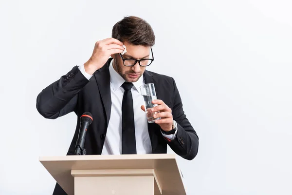 Asustado hombre de negocios en traje de pie en el tribuno podio y sosteniendo un vaso de agua durante la conferencia aislado en blanco - foto de stock