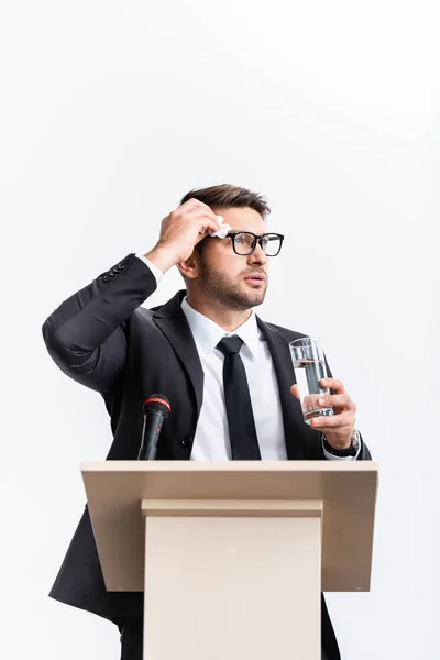 Spaventato uomo d'affari in giacca e cravatta in piedi sul podio tribuna e tenendo un bicchiere d'acqua durante la conferenza isolato su bianco — Foto stock