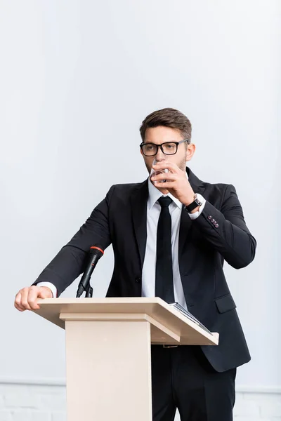 Scared businessman in suit standing at podium tribune and drinking water during conference isolated on white — Stock Photo