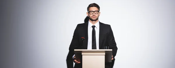 Панорамний знімок бізнесмена в костюмі, що стоїть на трибуні подіуму і дивиться на камеру під час конференції на білому тлі — стокове фото