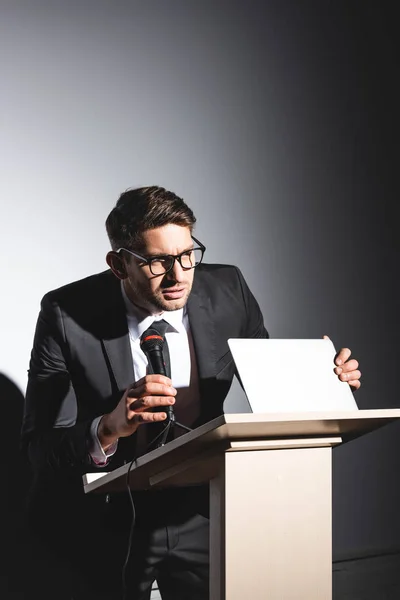 Spaventato uomo d'affari in giacca e cravatta in piedi sul podio tribune e tenendo il computer portatile durante la conferenza su sfondo bianco — Foto stock