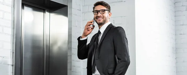 Панорамный снимок улыбающегося бизнесмена в формальной одежде разговаривающего на смартфоне возле лифта — стоковое фото