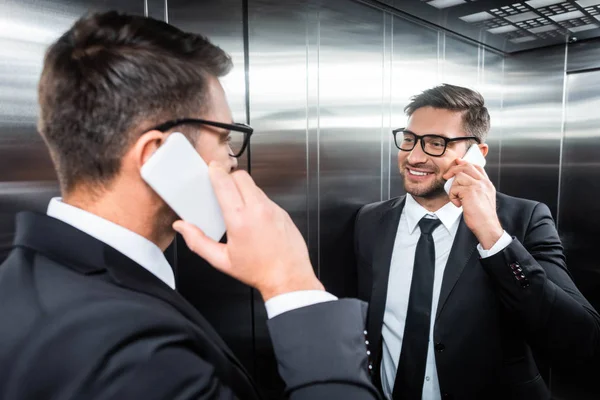 Smiling businessman in suit talking on smartphone and looking at mirror in elevator — Stock Photo