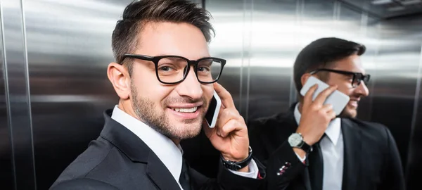 Panoramic shot of smiling businessman in suit talking on smartphone in elevator — Stock Photo