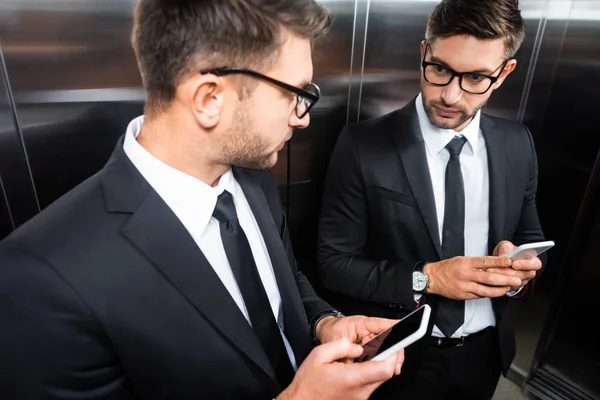 Selective focus of businessman in suit holding smartphone and looking at mirror in elevator — Stock Photo