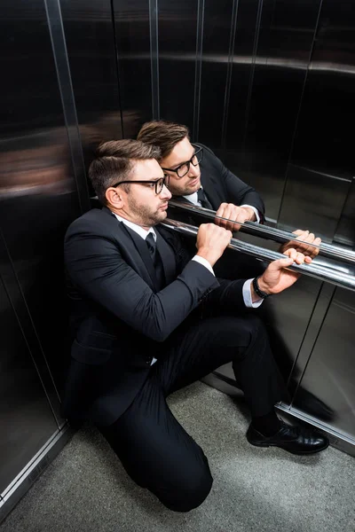 Vista de ángulo alto del hombre de negocios asustado en traje con claustrofobia sentado en el piso en ascensor - foto de stock