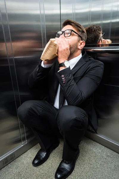 Hombre de negocios asustado en traje con claustrofobia respirando en bolsa de papel en ascensor - foto de stock