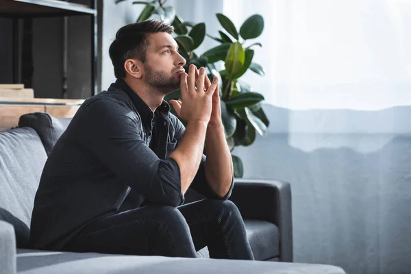 Vue latérale de l'homme beau et cher assis sur le canapé et regardant vers le haut dans l'appartement — Photo de stock