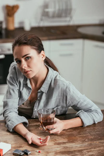 Frau blickt in Kamera, während sie Whiskeyglas und Pillen in der Hand hält — Stockfoto