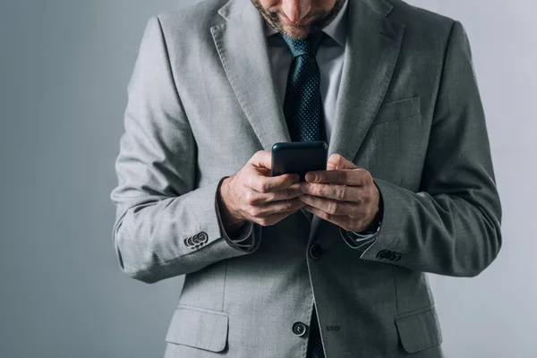 Обрезанный вид человека в костюме, держащего смартфон на сером фоне — стоковое фото