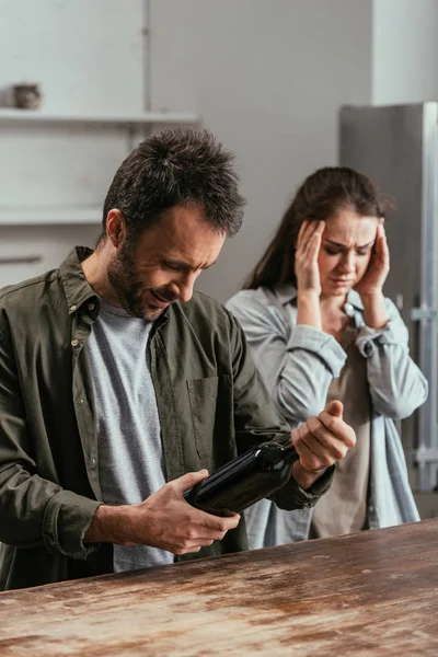 Foco seletivo de viciado em álcool homem segurando garrafa de vinho ao lado da esposa preocupada — Fotografia de Stock