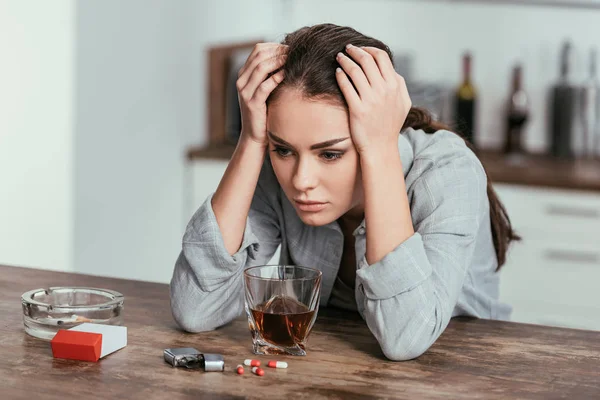 Enttäuschte Frau sitzt mit Whisky, Tabletten und Zigaretten am Tisch — Stockfoto