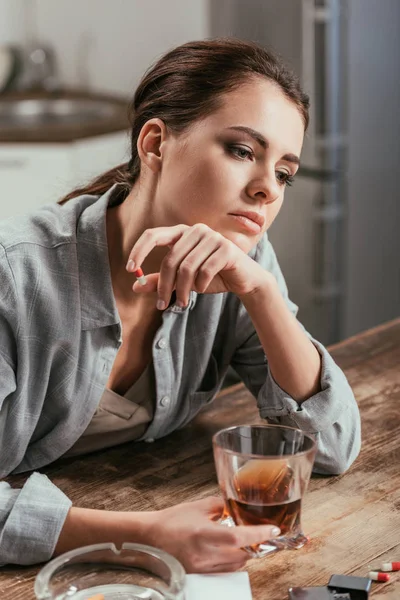 Mujer preocupada con adicción alcohólica sosteniendo vaso de whisky en la mesa - foto de stock