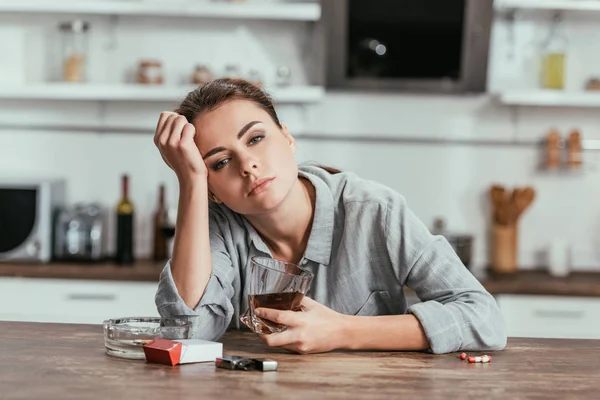 Traurigkeit Frau mit Alkoholsucht hält Whisky-Glas neben Tabletten und Zigaretten auf Tisch — Stockfoto