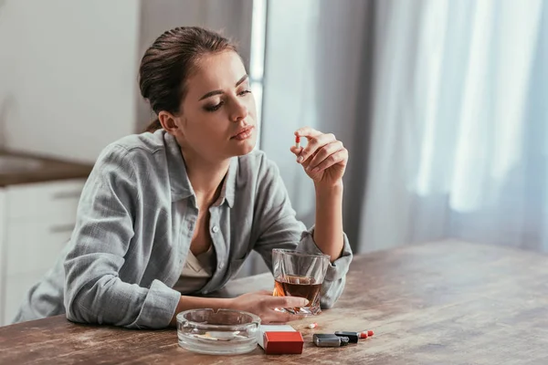 Femme alcoolique tenant pilule et verre de whisky à côté des cigarettes sur la table — Photo de stock