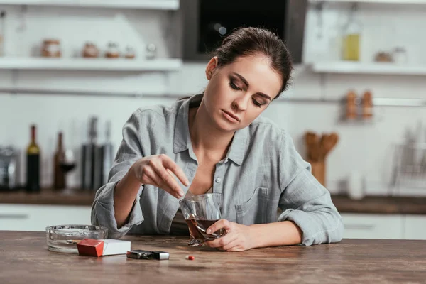 Donna alcolizzata che tiene il bicchiere di whisky accanto alle sigarette sul tavolo della cucina — Foto stock