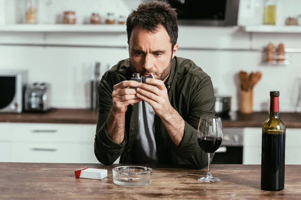 Worried man holding lighter beside wine and cigarettes on kitchen table — Stock Photo