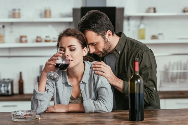 Homem perturbado abraçando álcool viciado esposa com copo de vinho na cozinha — Fotografia de Stock