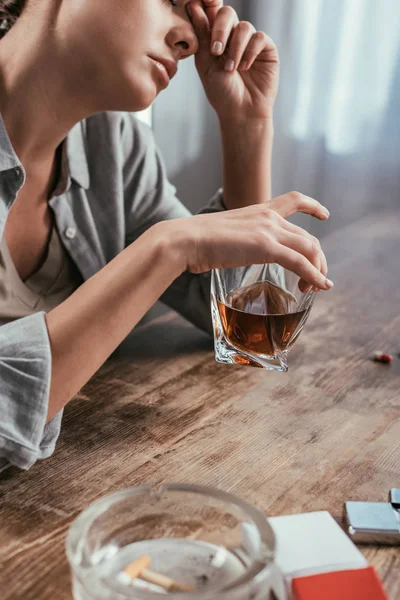 Enfoque selectivo de la mujer con adicción al alcohol sosteniendo vaso de whisky junto a los cigarrillos en la mesa - foto de stock
