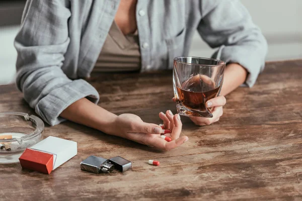 Vista ritagliata di donna che tiene bicchiere di whisky e pillole accanto alle sigarette sul tavolo — Foto stock
