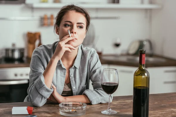 Mulher pensativa fumando ao lado de vinho na mesa da cozinha — Fotografia de Stock