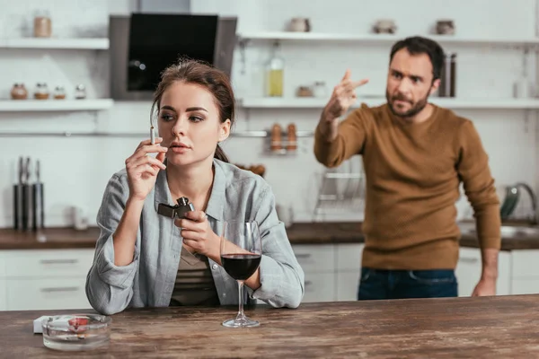 Foco seletivo da mulher com o vinho fumando enquanto marido irritado mostrando o dedo médio — Fotografia de Stock