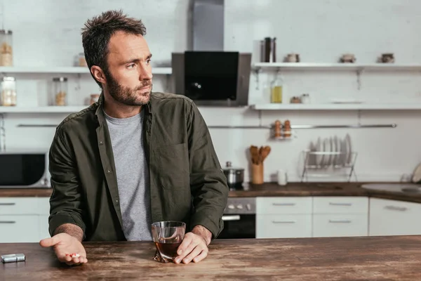 Trauriger Mann hält Whiskey-Glas und Pillen auf Küchentisch — Stockfoto