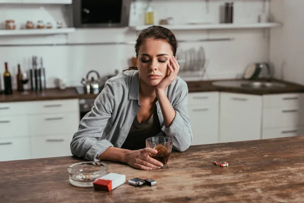 Беспокойная женщина держит стакан виски рядом с сигаретами и таблетками на столе — стоковое фото