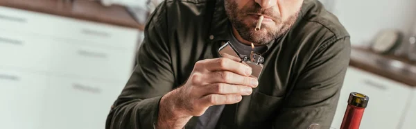 Обрезанный вид человека, зажигающего сигарету рядом с бутылкой вина, панорамный снимок — стоковое фото
