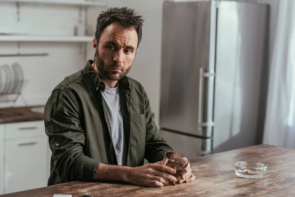 Worried man holding whiskey glass beside ashtray on kitchen table — Stock Photo