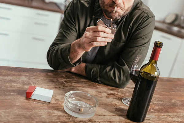 Vista cortada do homem acendendo cigarro ao lado de garrafa de vinho e vidro na mesa — Fotografia de Stock