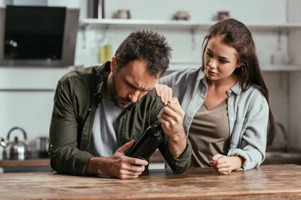 Mujer apoyando la adicción al alcohol marido con botella de vino en la cocina - foto de stock