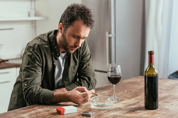 Hombre decepcionado fumando cigarrillo junto a la botella de vino y vidrio en la mesa de la cocina - foto de stock