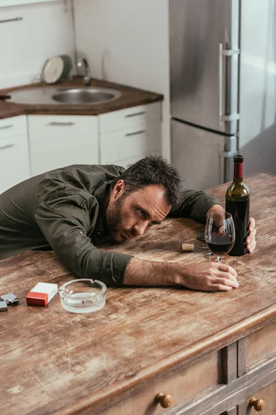 Hombre borracho sosteniendo botella de vino y vidrio junto a cigarrillos en la mesa de la cocina - foto de stock