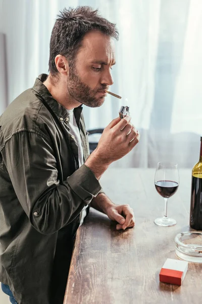 Hombre iluminación cigarrillo al lado de botella de vino y vidrio en la mesa - foto de stock