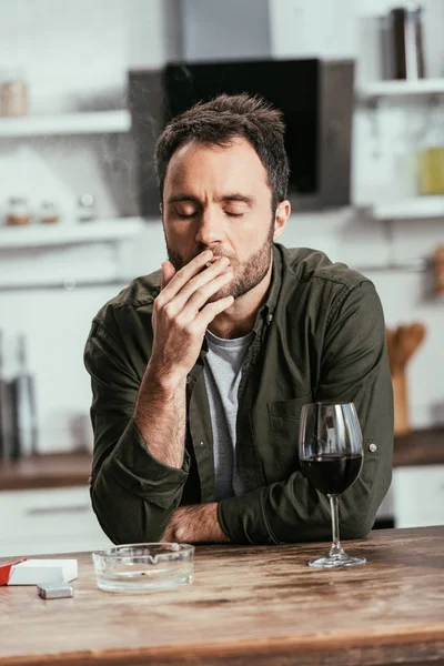 Мужчина курит сигарету рядом с бокалом вина на кухонном столе — стоковое фото