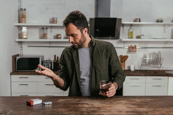 Uomo con bicchiere di whisky in possesso di pillole in cucina — Foto stock