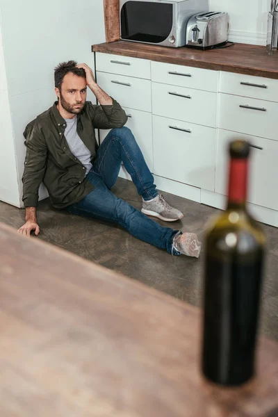 Селективный фокус человека с алкогольной зависимостью, сидящего на полу и смотрящего на бутылку вина на кухонном столе — стоковое фото