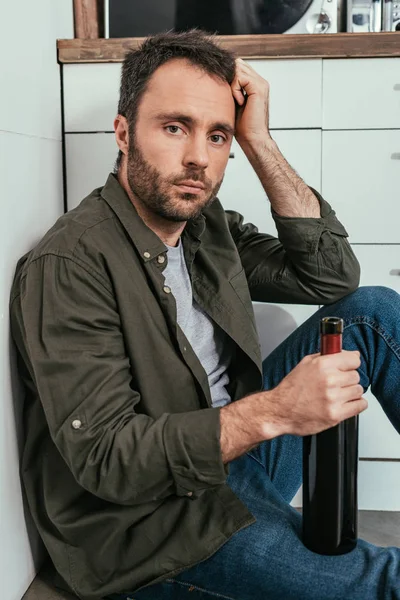 Thoughtful man holding wine bottle and looking at camera on kitchen floor — Stock Photo