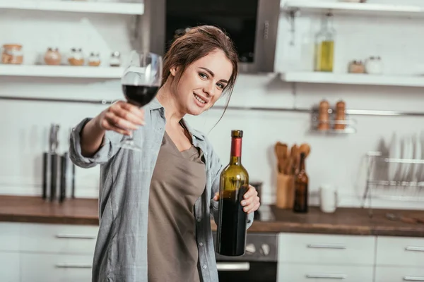 Foco seletivo de mulher sorridente segurando garrafa e copo de vinho na cozinha — Fotografia de Stock