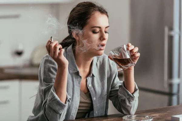 Mujer fumando y mirando el vaso de whisky en la mesa de la cocina - foto de stock