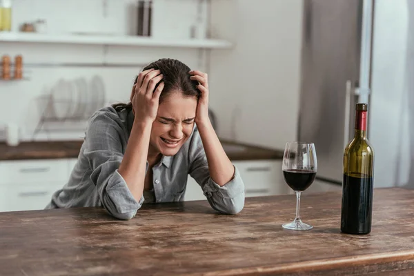Женщина плачет возле бокала вина и бутылки на кухонном столе — стоковое фото