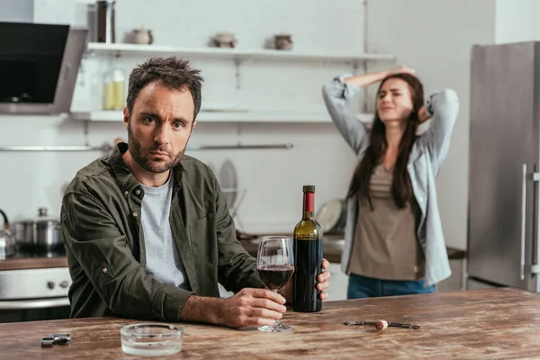 Foco seletivo do homem viciado em álcool com vinho e esposa chateada na cozinha — Fotografia de Stock